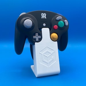 Könnte beinhalten: Ein schwarzer GameCube-Controller mit einem weißen Ständer. Der Ständer hat ein 3D-gedrucktes Design mit einem Würfel und dem Buchstaben G.