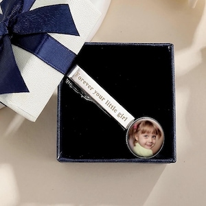 Könnte beinhalten: Eine silberne Krawattenklammer mit einem Foto eines kleinen Mädchens und dem eingravierten Text "Forever your little girl". Die Krawattenklammer wird in einer schwarzen Schachtel mit einer weißen und marineblauen Geschenkbox und Schleife präsentiert.