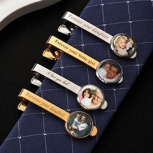 May include: Four metal tie clips in silver and gold tones, each featuring a circular photo insert and engraved text. The clips display phrases including Forever your daughter, Forever your little girl, and I love you dad, alongside various family photographs.