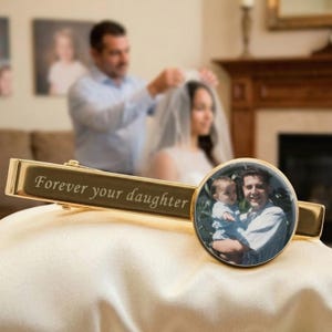 May include: A gold-colored tie clip with the engraved text "Forever your daughter." The clip features a circular photo of a man holding a baby. The background shows a wedding scene.