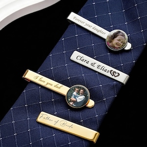 May include: Four personalized tie clips on a blue patterned tie. One gold clip reads "I love you dad" with a photo. Another silver clip says "Forever your daughter" with a photo. Other clips say "Clara & Elias" and "Father of Bride".
