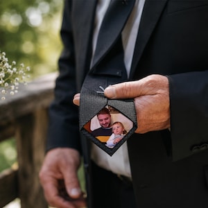 May include: A black necktie featuring a hidden interior pocket at the tip that reveals a rectangular color photograph of a man and a young child. The tie is made of a textured black fabric and is worn over a white dress shirt and black suit jacket.