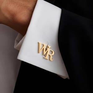 Könnte beinhalten: Ein goldfarbener Manschettenknopf mit den Buchstaben WR in einer Serifenschrift, befestigt an der weißen Manschette eines langärmeligen Hemdes. Der Manschettenknopf ist horizontal auf dem Stoff platziert und hat eine polierte Oberfläche.