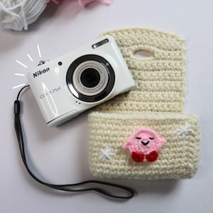 May include: A white Nikon Coolpix digital camera rests on a cream-colored crocheted camera case. The case features a pink character with red feet and white star accents. Yarn in white and pink is in the background.