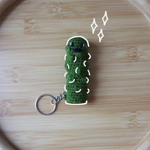 Può includere: Un portachiavi a forma di cetriolo verde all'uncinetto fatto a mano, con un volto sorridente e dettagli bianchi. Il portachiavi ha un anello in metallo argentato. Il cetriolo misura circa 7.6 cm di altezza.