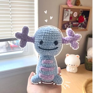 May include: A handmade crochet plush toy in the shape of a cartoon character. The plush is primarily light blue with lavender accents and features a smiling face and cross-shaped appendages. The toy is held in a person's hand.