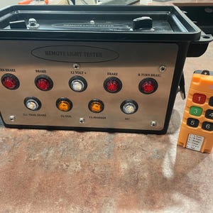 Puede incluir: Un comprobador de luces remoto negro y plateado con múltiples luces indicadoras y un mando a distancia naranja. El comprobador tiene etiquetas para varias funciones de luz, incluyendo freno e intermitentes. El mando a distancia tiene botones numerados.
