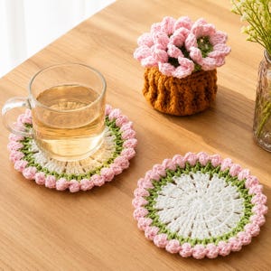 May include: Two round crochet coasters featuring a white center, a green middle ring, and a scalloped pink border. A clear glass mug filled with light-colored liquid sits on one coaster, and a small brown crochet pot with pink flowers sits nearby.