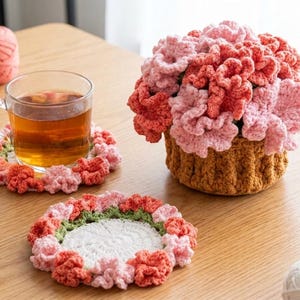 Pode incluir: Uma caneca de vidro transparente com chá está sobre um descanso de copo de crochê com detalhes de flores rosa e laranja. Um arranjo de flores de crochê em uma cesta marrom e dois descansos de copo adicionais também estão em uma superfície de madeira.