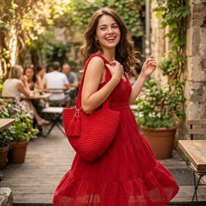 May include: A woman in a red dress carries a matching red crocheted tote bag with a tassel. The bag is held over the shoulder. The woman is smiling and walking on a wooden deck. The background includes a cafe with people and greenery.