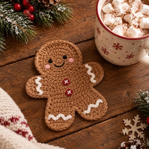 May include: A brown crochet gingerbread man coaster featuring a smiling face with pink cheeks, two red buttons on the chest, and white wavy icing details on the arms and legs. The coaster is placed on a dark wooden surface next to a mug.