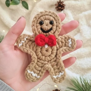May include: A tan crochet gingerbread man ornament featuring a bright red yarn bow tie, black plastic bead eyes, a stitched black smile, and white yarn accents on the arms and legs to resemble icing details.