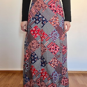 Peut inclure: Longue jupe patchwork avec un mélange de carrés à motifs rouges, bleus et marron. La jupe a une taille haute et une coupe fluide. Le haut est un t-shirt noir à manches longues.