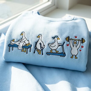 May include: Light blue sweatshirt featuring a whimsical design of cartoon ducks engaged in various fitness activities. The ducks are depicted on a massage table, using crutches, running on a treadmill, and lifting weights. Red heart accents are included.