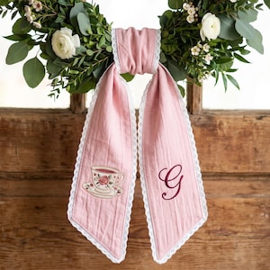 Pode incluir: Uma fita de tecido rosa empoeirado com acabamento em renda branca nas bordas. Um lado exibe uma xícara de chá bordada com um padrão floral, enquanto o outro lado apresenta uma letra G bordada em rosa escuro em fonte cursiva.