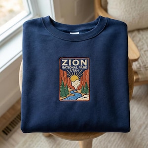 Puede incluir: Sudadera azul marino con un parche rectangular con las palabras "ZION NATIONAL PARK UTAH". El parche representa un paisaje con un río, montañas, árboles y un sol. La sudadera está doblada sobre una silla de madera.