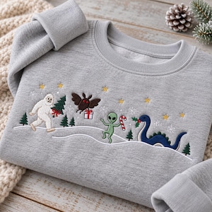 May include: A heather gray crewneck sweatshirt featuring embroidered holiday designs including a white yeti holding a tree, a brown moth with a gift, a green alien with a candy cane, and a blue sea monster, all set against a snowy landscape.