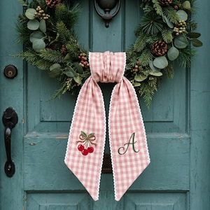 May include: A pink and white gingham fabric ribbon with white lace trim, featuring an embroidered red cherry motif on one tail and the letter A in green script on the other, hanging from a green pine wreath with pinecones and eucalyptus.