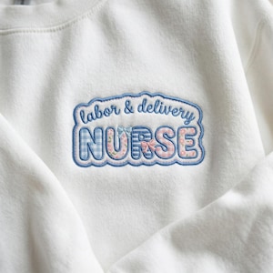 Puede incluir: Sudadera blanca con un diseño bordado. El diseño presenta las palabras "labor & delivery NURSE" en un esquema de color azul y rosa. La palabra "NURSE" tiene un patrón de cuadros y detalles florales.