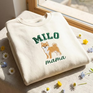 Op de afbeelding: Crèmekleurige sweatshirt met de naam "MILO" in groene blokletters boven een geborduurde Shiba Inu hond. Het woord "mama" is eronder geborduurd in groen. De sweatshirt is opgevouwen, met bloemen eromheen.