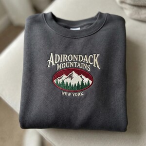 May include: A dark gray sweatshirt with embroidered text and a mountain scene. The text reads "ADIRONDACK MOUNTAINS NEW YORK" in cream-colored thread. The mountain scene is in a red oval with white mountains and green trees.
