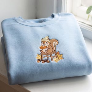 May include: A light blue crewneck sweatshirt featuring an embroidered design of a brown squirrel wearing a cream-colored knit hat, sitting on a wooden chair while eating a slice of pumpkin pie, surrounded by fallen autumn leaves and acorns.