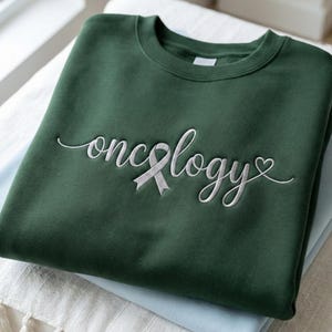 May include: A dark green sweatshirt with the word "oncology" embroidered in white script, featuring a white ribbon and a heart symbol. The sweatshirt is folded on top of other folded items.