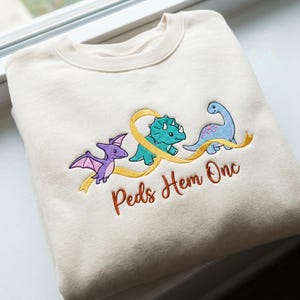 Może przedstawiać: Kremowa bluza z haftowanymi dinozaurami i żółtą wstążką. Projekt zawiera fioletowego Pterodaktyla, turkusowego Triceratopsa i niebieskiego Brontozaura. Tekst "Peds Hem Onc" jest haftowany na brązowo.