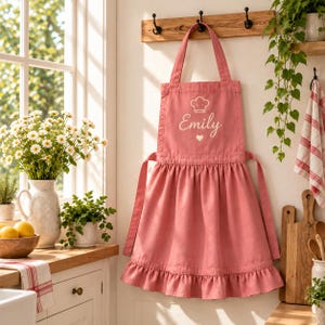 May include: A dusty rose-colored apron featuring a ruffled skirt, a neck loop, and waist ties. The chest area displays a white graphic of a chef hat, the name Emily in cursive script, and a small heart icon centered below the name.