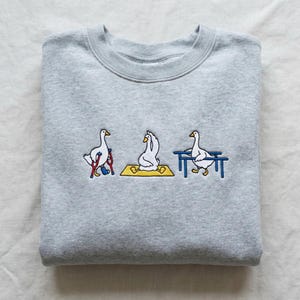 May include: A pale grey sweatshirt with an embroidered design of three geese. One goose is on crutches, another is in a yoga pose on a yellow mat, and the third is near a blue bench. The design is a humorous take on yoga.