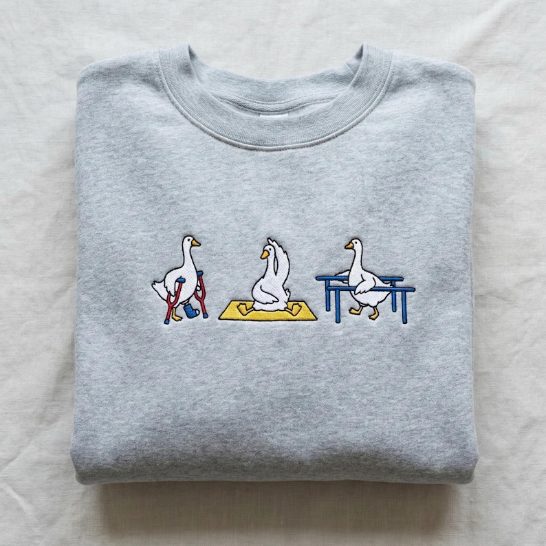 May include: A pale grey sweatshirt with an embroidered design of three geese. One goose is on crutches, another is in a yoga pose on a yellow mat, and the third is near a blue bench. The design is a humorous take on yoga.