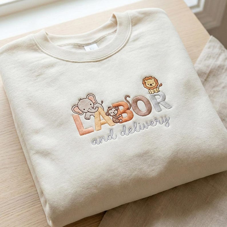Op de afbeelding: Cr&egrave;mekleurige sweatshirt met geborduurde tekst en dierlijke afbeeldingen. Het woord "LABOR" staat in oranje en bruine letters, met een olifant en een aap. De woorden "and delivery" staan in grijze letters, met een leeuw bovenop de "R".