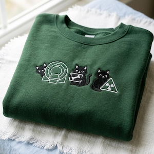 Könnte beinhalten: Ein dunkelgrünes Sweatshirt mit gestickten Designs von schwarzen Katzen und medizinischen Symbolen. Das Design umfasst eine Katze mit einem MRT-Gerät, eine Katze, die einen Röntgenknochen hält, und eine Katze mit einem Strahlungssymbol. Das Sweatshirt ist auf einer weißen und blauen Oberfläche gefaltet.