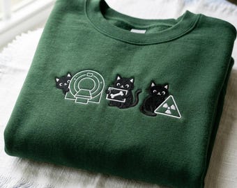 Op de afbeelding: Een donkergroene sweatshirt met geborduurde ontwerpen van zwarte katten en medische symbolen. Het ontwerp omvat een kat met een MRI-machine, een kat met een röntgenbot en een kat met een stralingssymbool. De sweatshirt is op een witte en blauwe ondergrond gevouwen.