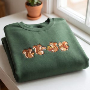May include: A forest green crewneck sweatshirt featuring an embroidered design of four brown squirrels. The squirrels are depicted holding an acorn, a small pie, a colourful autumn leaf, and wearing a plaid scarf, respectively.