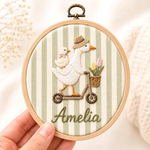 May include: A round wooden embroidery hoop containing fabric with vertical sage green and white stripes. The embroidery features a white goose wearing a straw hat on a scooter, carrying a small duck and a basket of yellow and pink tulips. The name Amelia is stitched in green cursive text below.