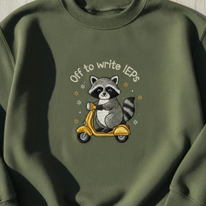 Könnte beinhalten: Olivgrünes Sweatshirt mit einem gestickten Design. Das Design zeigt einen Waschbären auf einem gelben Roller mit dem Text "Off to write IEPs". Kleine Sternakzente umgeben den Waschbären und den Text.