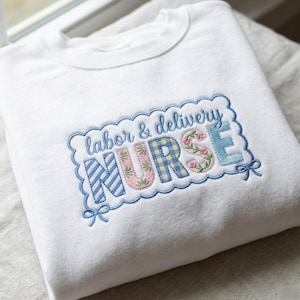 Puede incluir: Sudadera blanca con el texto bordado "labor & delivery NURSE" en un marco festoneado azul. La palabra "NURSE" presenta patrones florales y a cuadros en rosa, azul y amarillo. Lazos azules acentúan el marco.