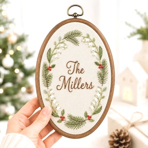 May include: An oval wooden embroidery hoop containing cream-colored fabric with a hand-stitched wreath of green pine needles, white lily of the valley flowers, and small red berries. The text "The Millers" is embroidered in brown script in the center.