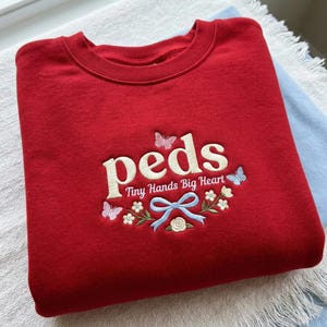 May include: A red crewneck sweatshirt with the embroidered word "peds" in cream-colored letters. Below the word are the words "Tiny Hands Big Heart" with floral and butterfly accents. A light blue bow is also embroidered.