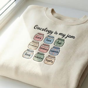 Peut inclure: Sweat-shirt crème avec l'inscription "Oncology is my jam" en noir. En dessous, neuf pots avec des mots comme "care", "love" et "fun" brodés. Les pots sont de différentes couleurs pastel.