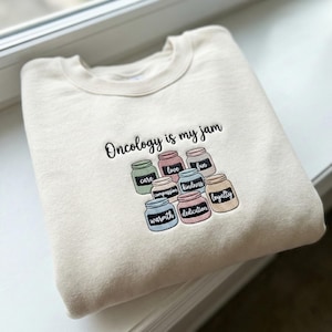 Peut inclure: Sweat-shirt crème brodé avec la phrase "Oncology is my jam". En dessous, des bocaux brodés avec des mots comme "care", "love" et "kindness". Le sweat-shirt est plié sur un rebord de fenêtre.