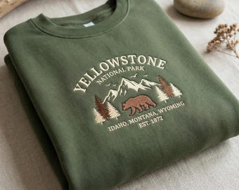 Op de afbeelding: Olijfgroene sweatshirt met geborduurd "Yellowstone National Park" ontwerp. Voorzien van een beer, bergen en bomen. Tekst: "Idaho, Montana, Wyoming, Est. 1872". Een comfortabel kledingstuk met natuurthema.