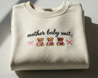 May include: Cream-colored sweatshirt with the embroidered text "mother baby unit". Below the text are three brown teddy bears with pink bows and pink bows on either side. The sweatshirt is folded neatly.