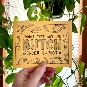 May include: A yellow card with black illustrations and the text "THINGS THAT GIVE ME BUTCH GENDER EUPHORIA" in gold glitter lettering. The card is held in front of a background of green plants.