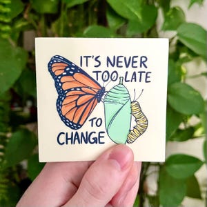May include: A square sticker with a monarch butterfly illustration in orange, black, and white, next to a green chrysalis and a yellow caterpillar. The text "IT'S NEVER TOO LATE TO CHANGE" is written in navy blue.