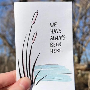 May include: A white card with black ink and a hand-drawn illustration of cattails in a pond. The text on the card reads "We have always been here."