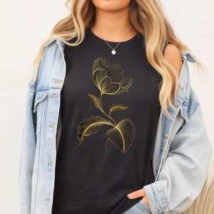 May include: Black t-shirt with a gold floral design. The graphic shows a stylized flower with detailed lines and leaves. The design is centered on the shirt, creating a contrast against the dark fabric. The shirt is a classic crew neck style.