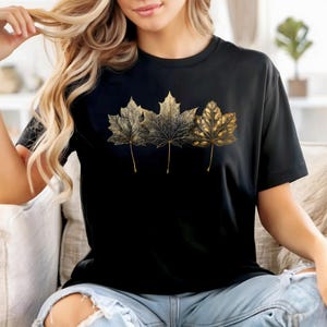 May include: Black t-shirt featuring three gold-colored maple leaves. The leaves are detailed with intricate veining and are arranged horizontally across the front of the shirt. The shirt has a classic crew neck and short sleeves.