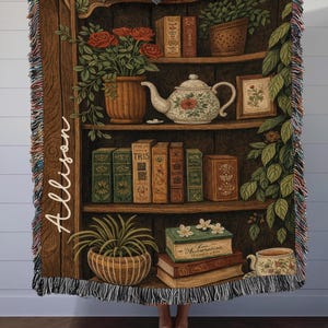 May include: A woven throw blanket featuring a bookshelf design with books, plants, and a teapot. The blanket has a brown background with the name "Allison" written vertically on the left side. The edges are fringed with multiple colors.